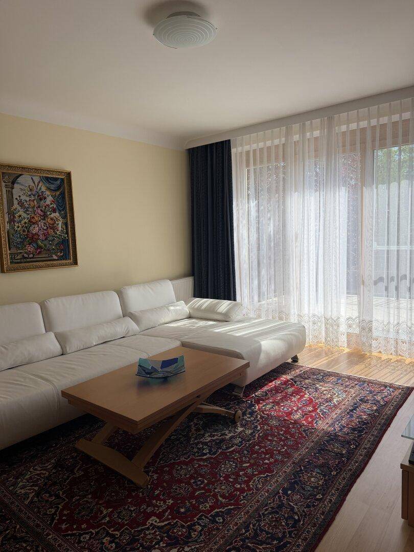 Helles Wohnzimmer mit weißem Ecksofa, Holztisch und großem Teppich vor der Terrassentür.