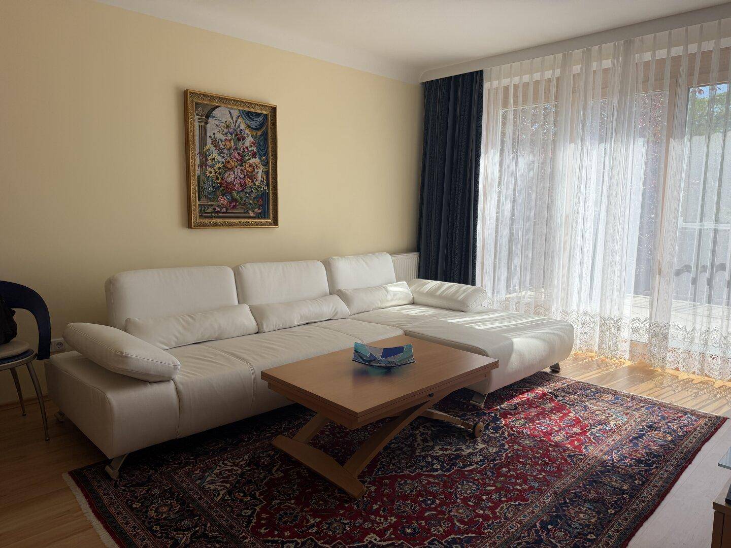 Geräumiges Wohnzimmer mit beigem Sofa, Holztisch und großem Fenster, das viel Licht hereinlässt.