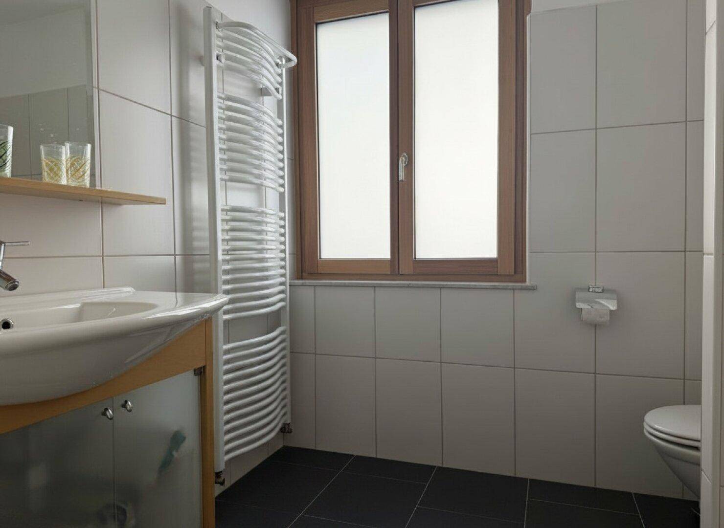 Helles Badezimmer mit WC, Waschbecken und beheiztem Handtuchhalter neben dem Fenster.