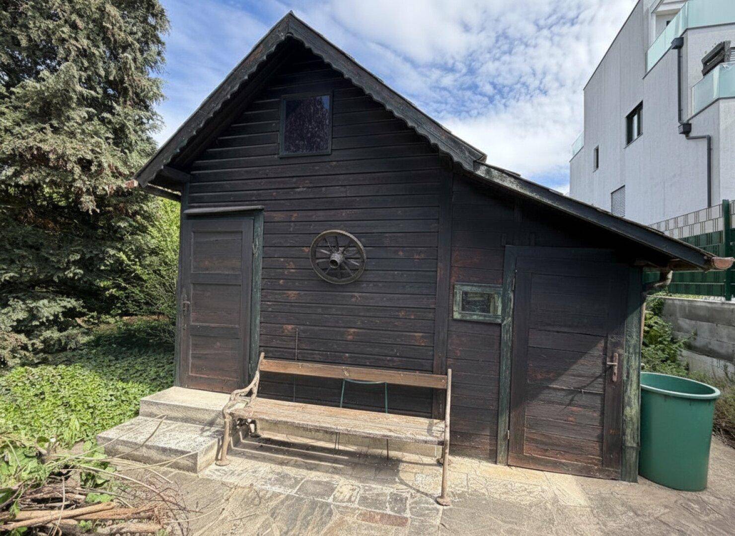 Rustikaler Holzschuppen mit einer alten Bank davor, umgeben von üppigem Grün im Garten.
