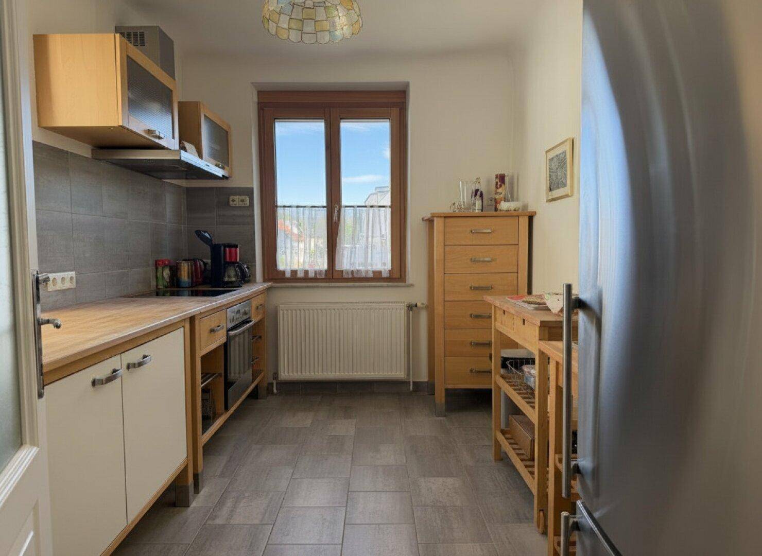 Funktionale Küche mit Holzfronten, Fliesenboden und Fenster mit Blick nach draußen.
