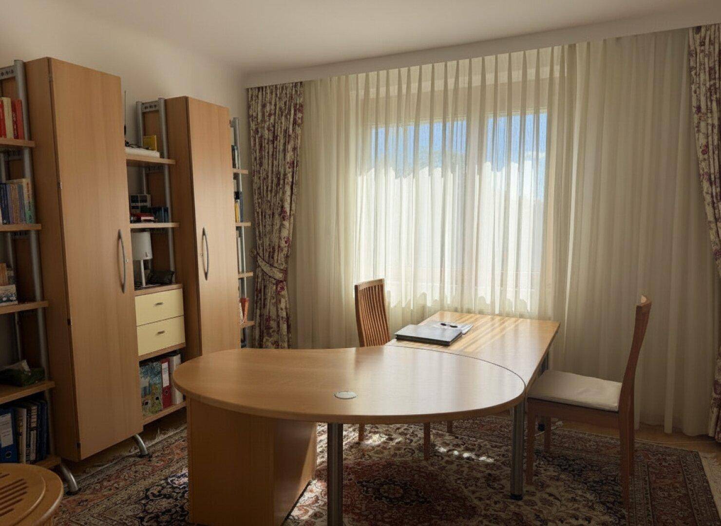 Helles Arbeitszimmer mit großem Holzschreibtisch, Bücherregalen und Teppichboden.