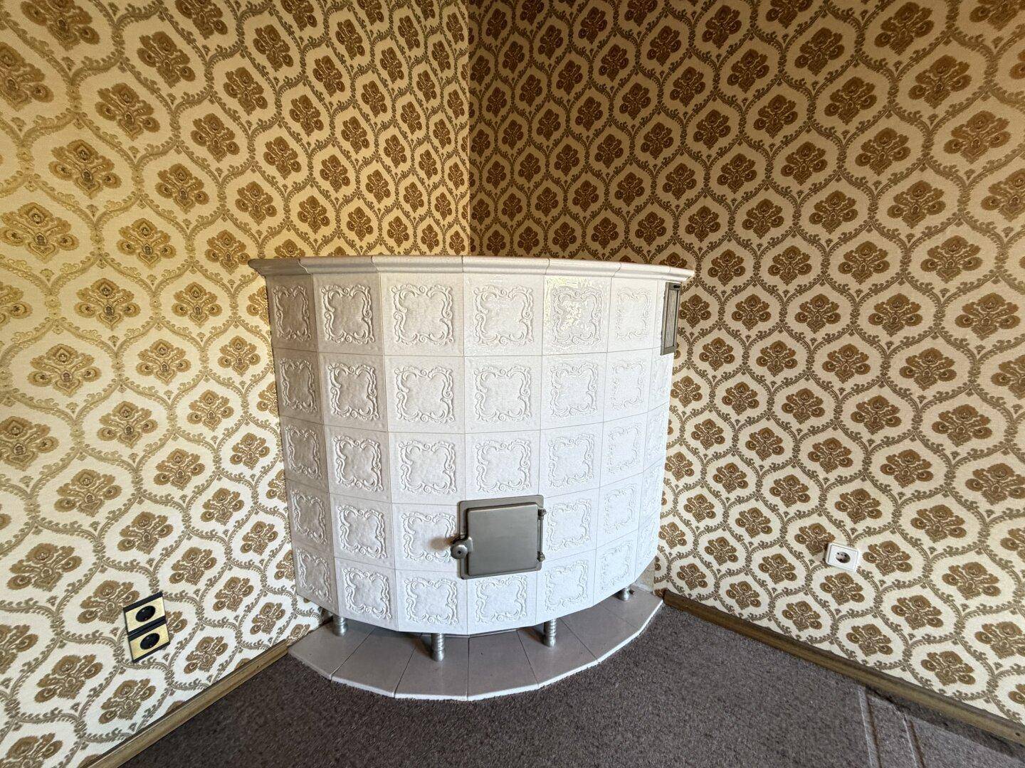 Weißer Kachelofen in einem Zimmer mit traditioneller gemusterter Tapete und Teppichboden.