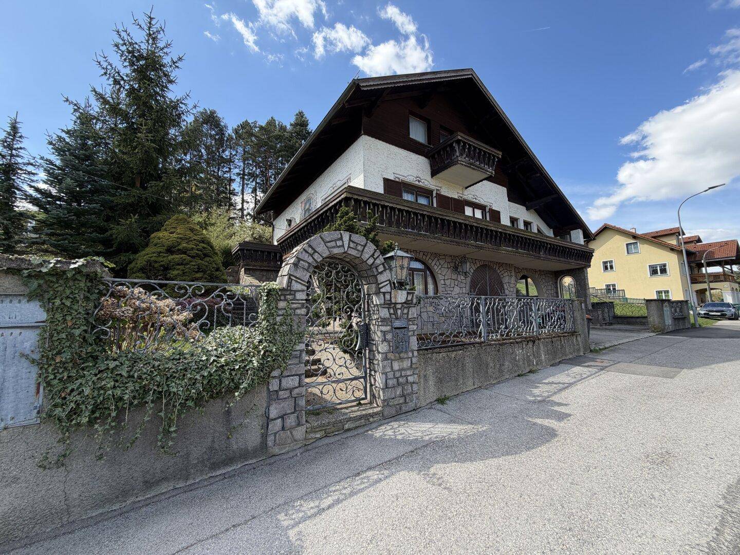 Traditionelles Haus mit Steinfassade, schmiedeeisernem Tor und Holzbalkonen an einer ruhigen Straße.