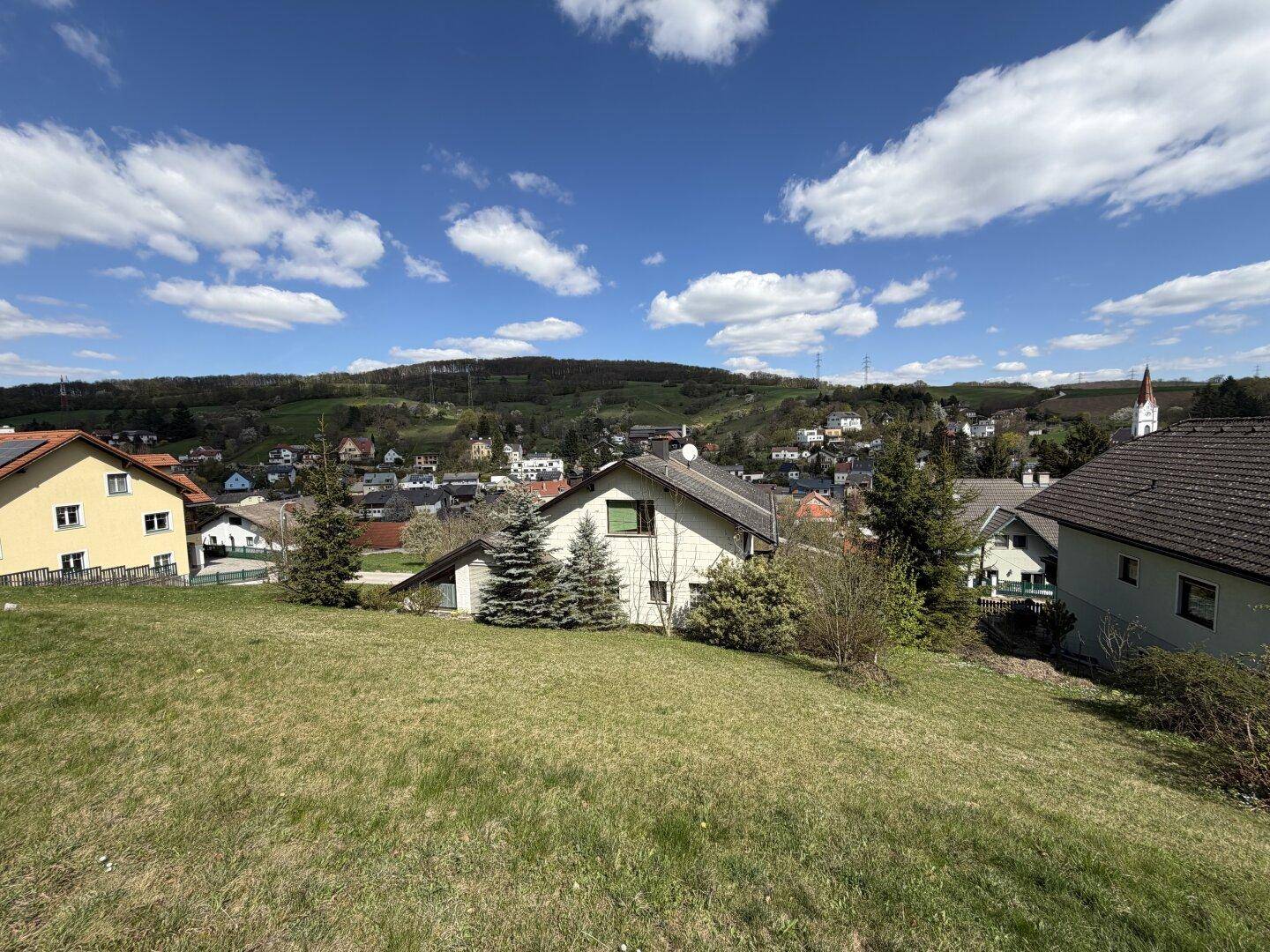 Grüne Rasenfläche mit Blick auf das Dorf und die umliegenden Hügel unter einem bewölkten Himmel.
