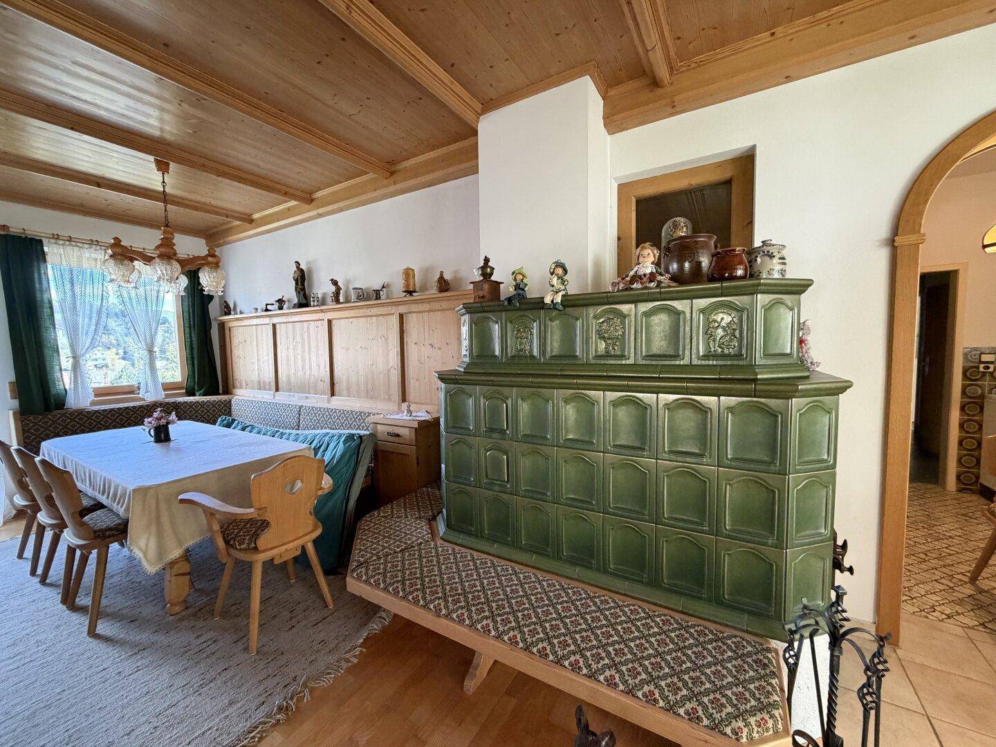 Traditioneller Wohn-Essbereich mit grünem Kachelofen, Holzbalkendecke und rustikalen Holzmöbeln.