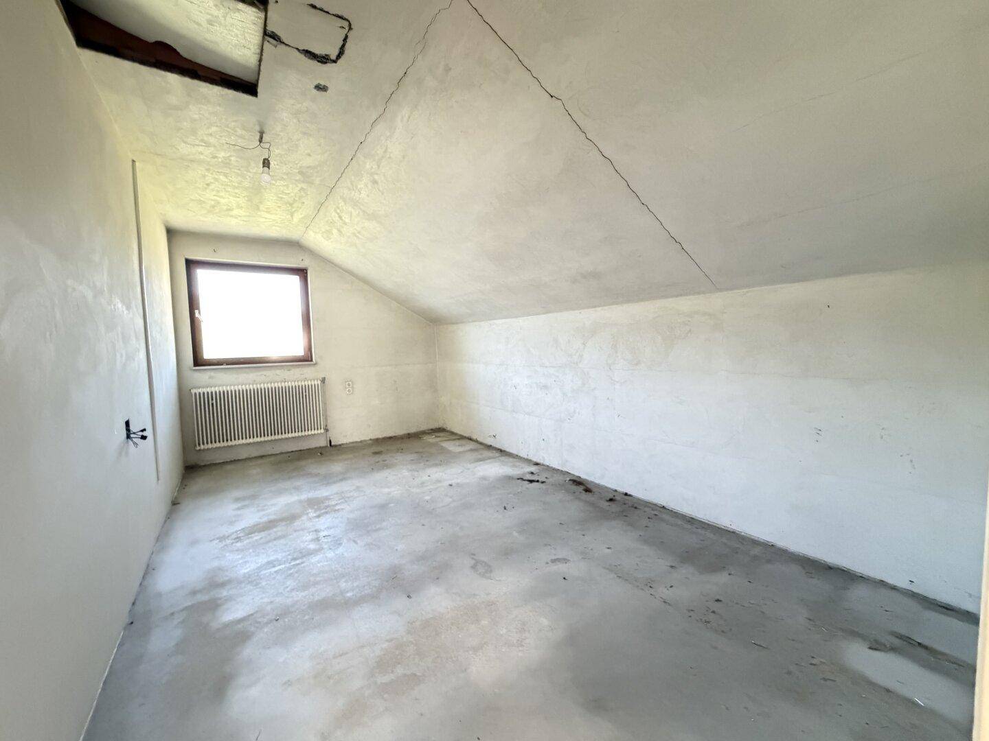 Unausgebauter Dachbodenraum mit schrägen Wänden und einem Fenster, bereit für den Ausbau.