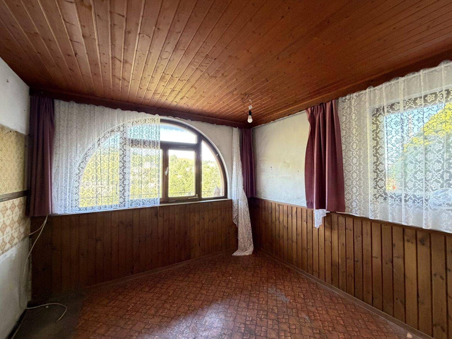 Zimmer mit Holztäfelung, Holzbalkendecke und einem markanten Rundbogenfenster.
