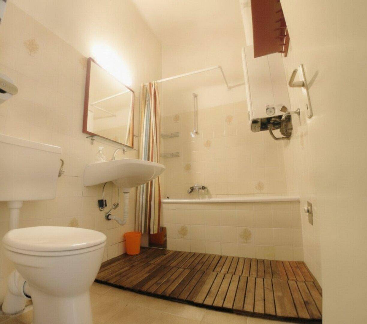 Badezimmer mit Badewanne, WC und Waschbecken, ausgestattet mit Fliesen und Holzrost.