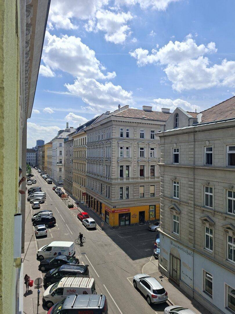 Stadtansicht mit Gebäuden, parkenden Autos und belebter Straße unter blauem Himmel.