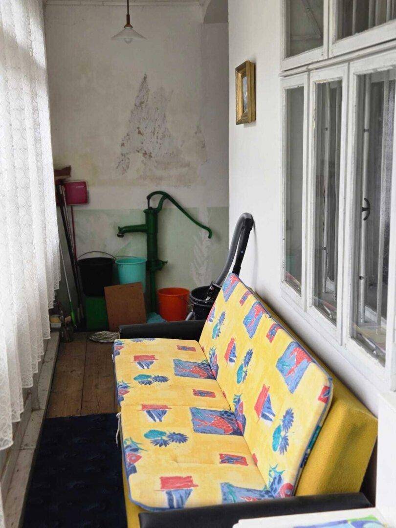 Verglaster Anbau mit bunter Couch, Holzboden und einer alten Wasserpumpe.