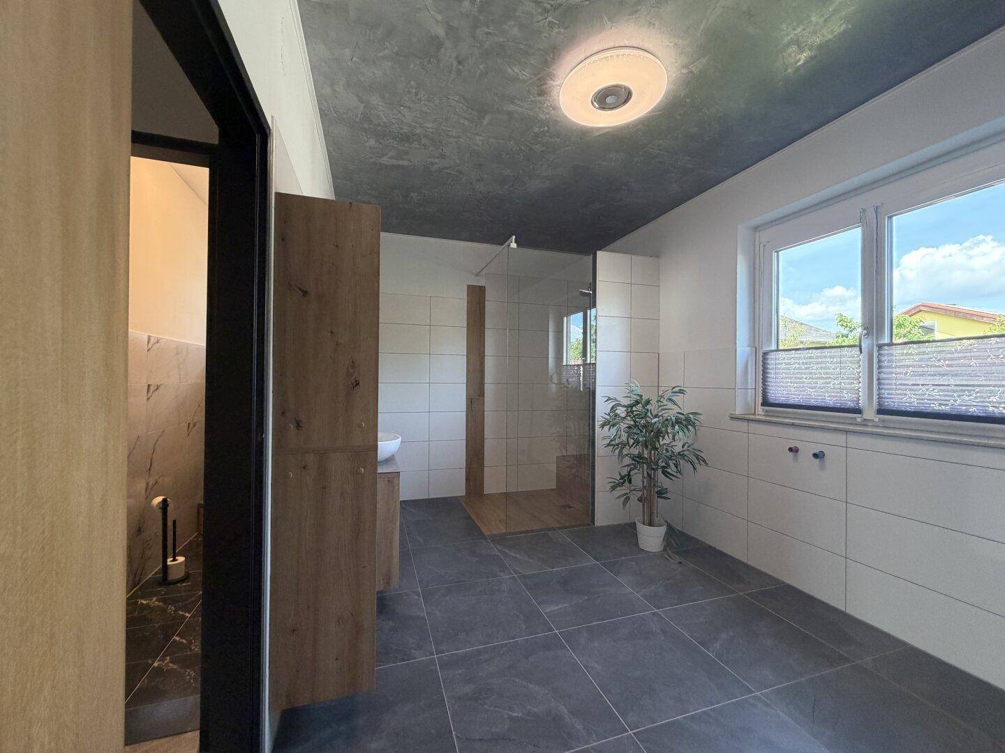 Geräumiges Badezimmer mit begehbarer Dusche, dunklen Fliesen und Fenster für Tageslicht.