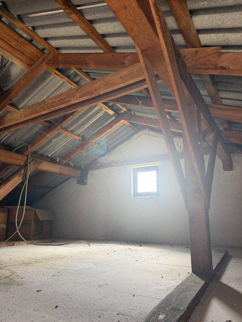 Unausgebauter Dachboden mit sichtbaren Holzbalken und einem kleinen Fenster, bietet Ausbaupotenzial.