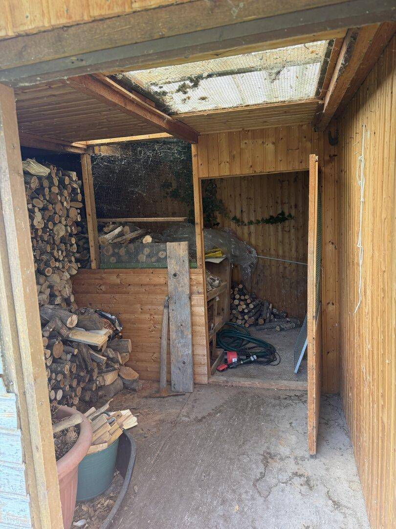 Innenansicht eines Holzschuppens mit gestapeltem Brennholz und Gartengeräten.
