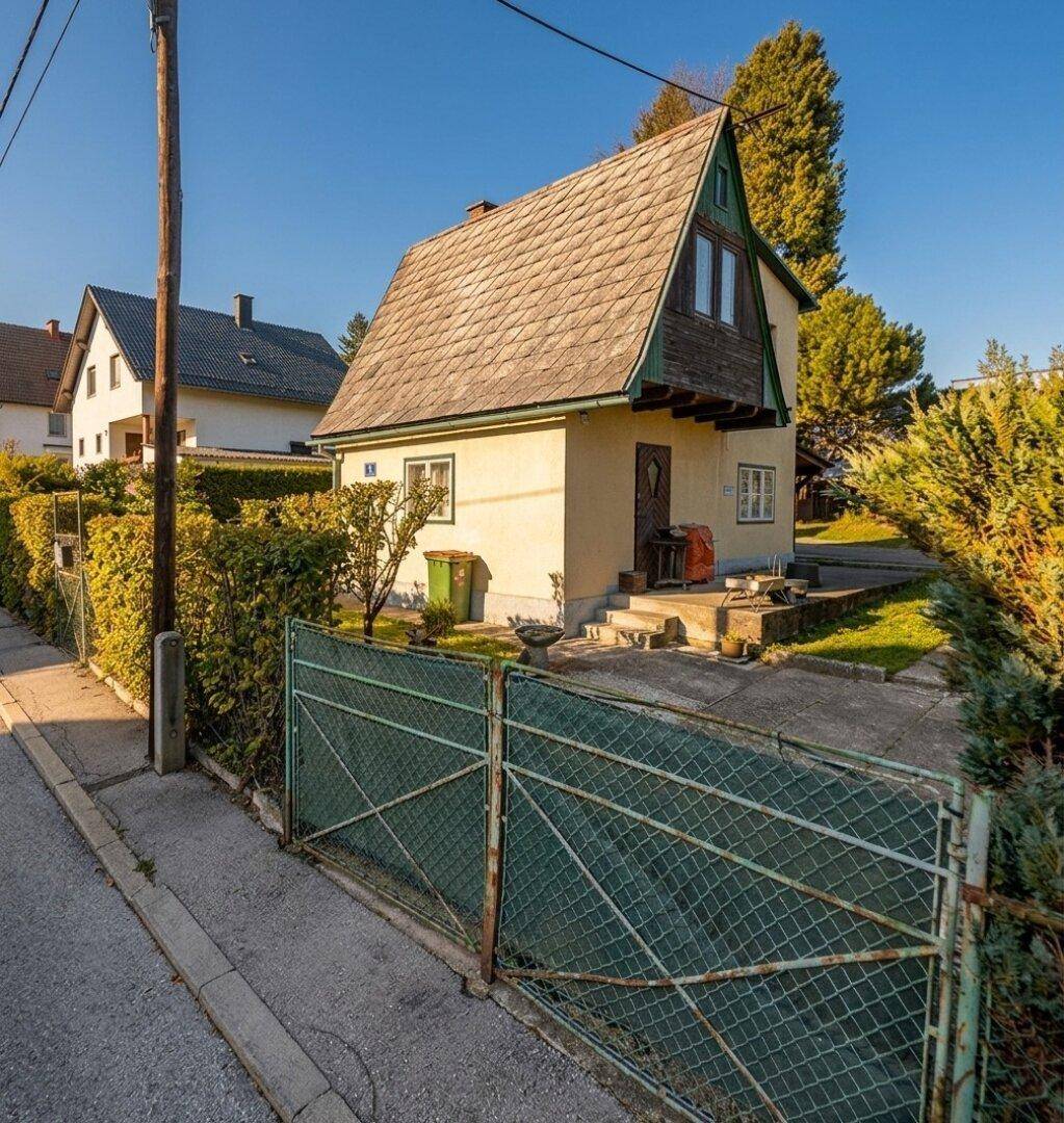 Seitenansicht des Hauses mit schrägem Dach und einfacher Fassade, umgeben von Hecken.