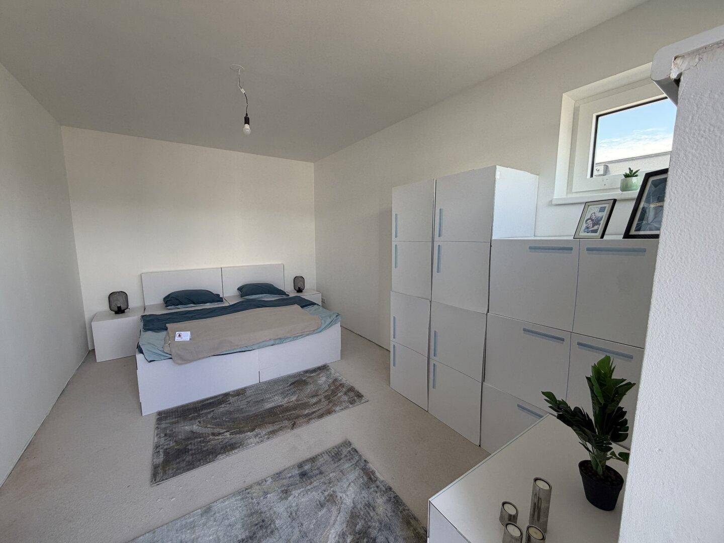 Schlafzimmer mit Fenster, modernem Bett und weißen Aufbewahrungsmöbeln, hell und funktional eingerichtet.