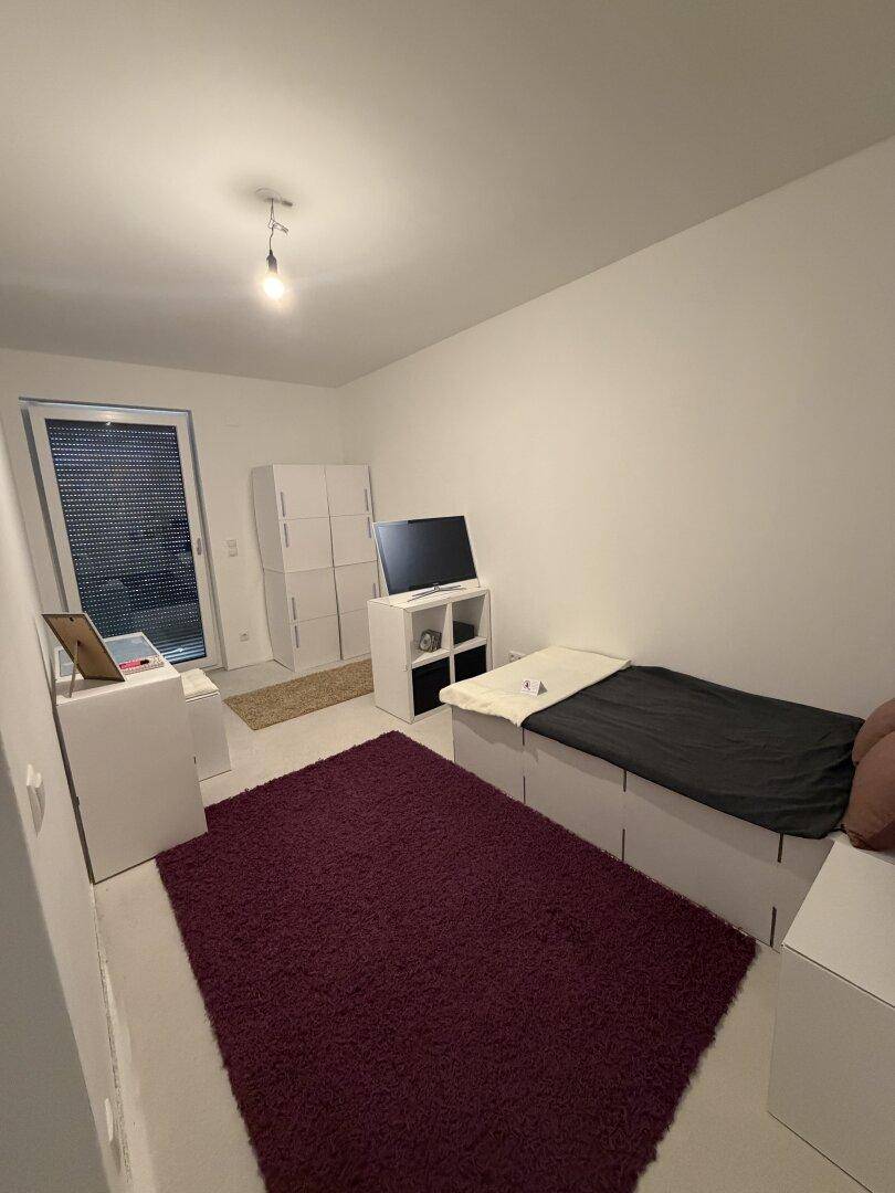 Kleineres Schlafzimmer mit Einzelbett, Fernseher und Zugang zu einem Balkon oder Garten, praktisch gestaltet.