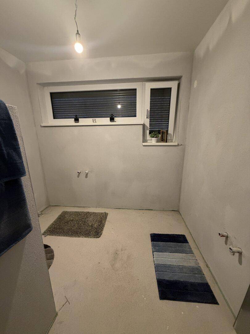Badezimmer im Rohbau mit zwei Fenstern und vorbereiteten Anschlüssen für Sanitäranlagen, bereit zum Ausbau.