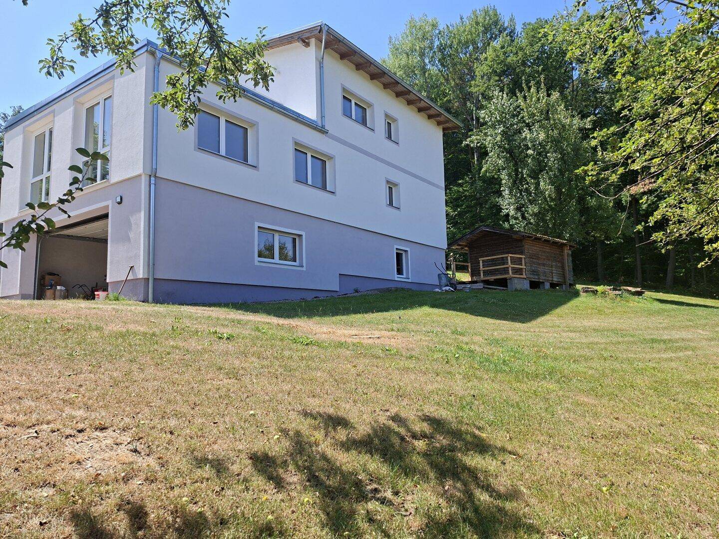 Rückansicht des Hauses mit großem Gartenbereich und einem kleinen Holzschuppen am Waldrand.