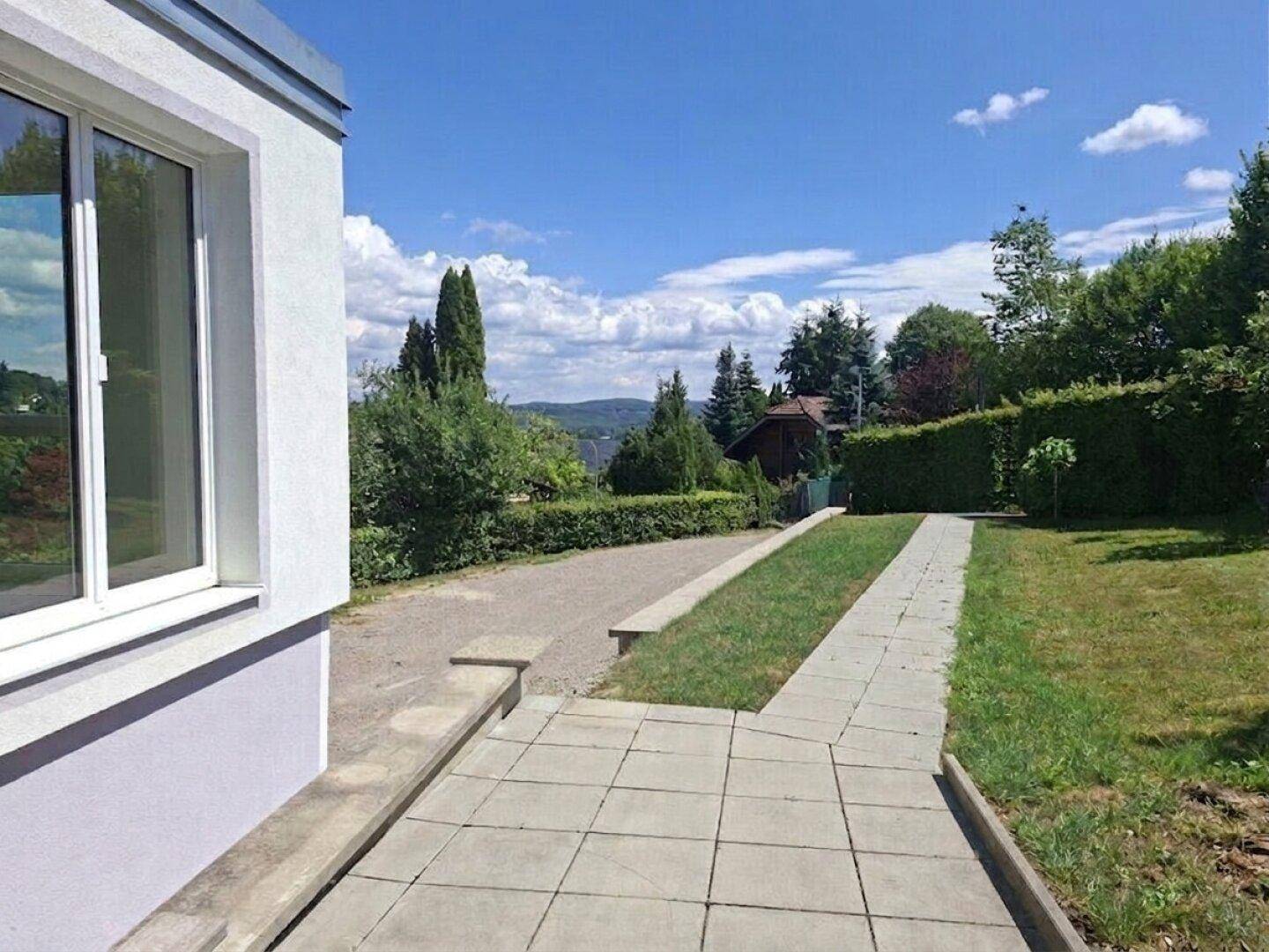 Gepflasterte Auffahrt zum Haus, umgeben von gepflegten Hecken und Bäumen unter blauem Himmel.