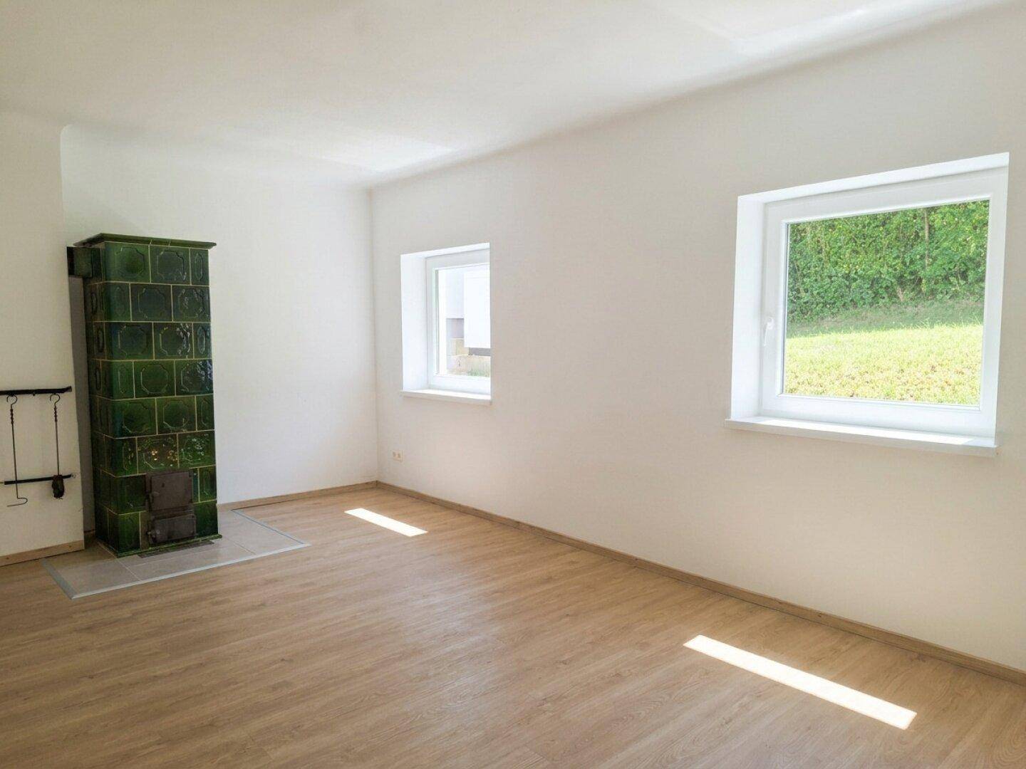 Heller Wohnraum mit traditionellem Kachelofen, Laminatboden und Fenstern mit Blick ins Grüne.