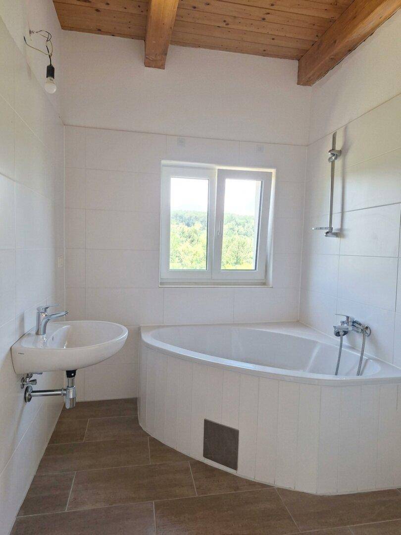 Badezimmer mit Eckbadewanne, Dusche, Waschbecken und Fenster mit Blick ins Grüne.