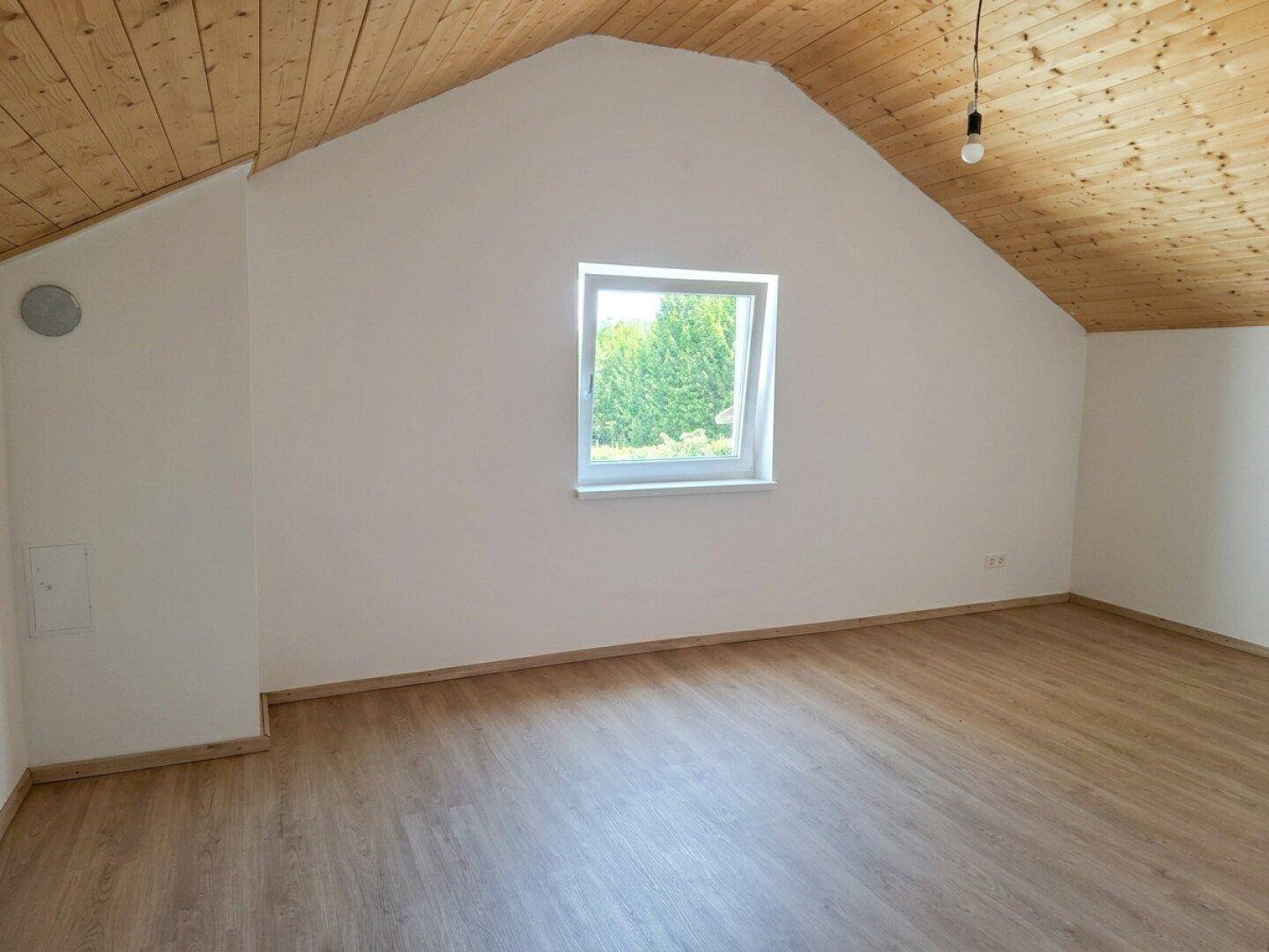Schlafzimmer im Dachgeschoss mit schräger Decke, Holzbalken und einem Dachfenster mit Blick ins Grüne.