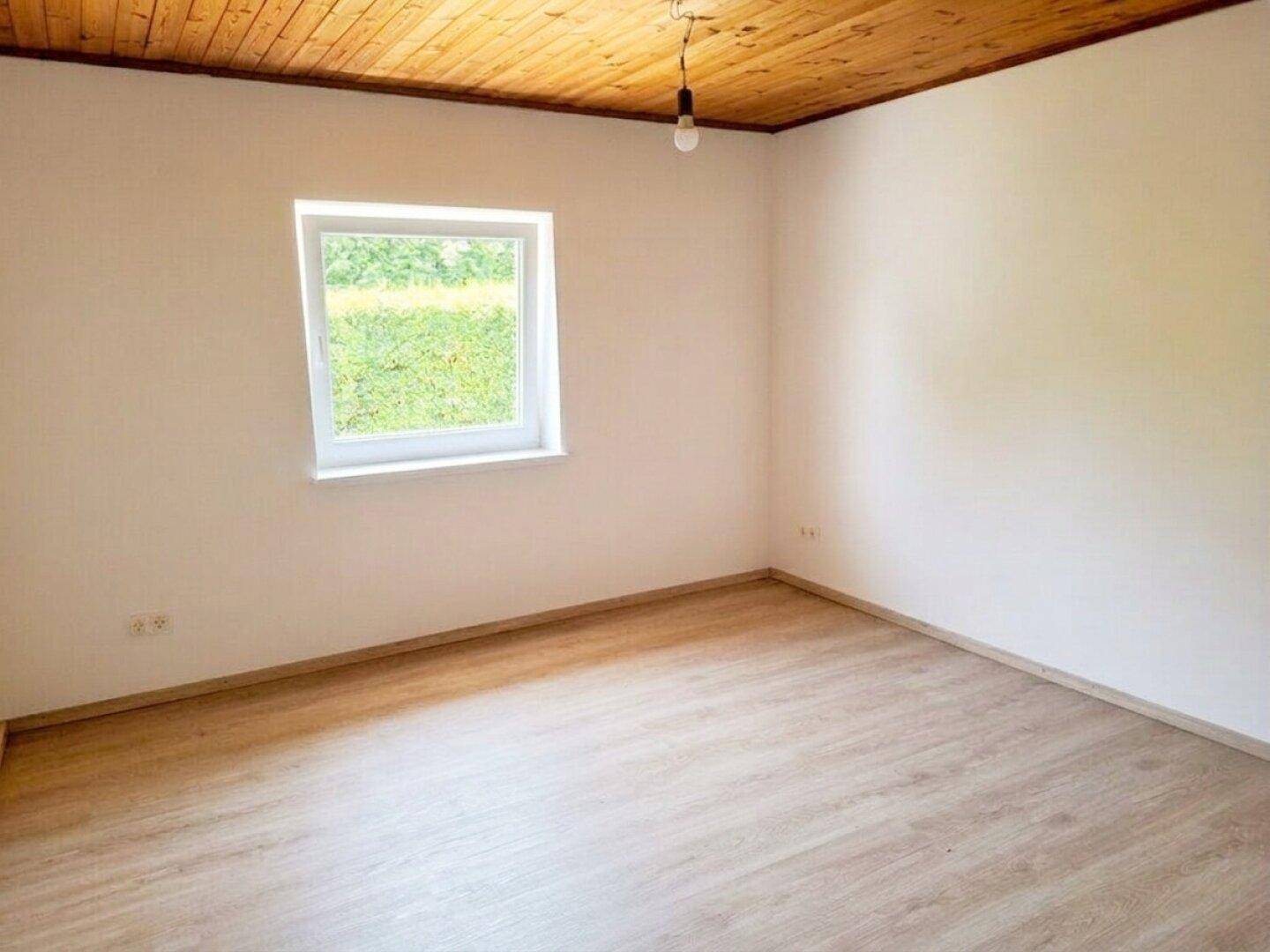 Helles Schlafzimmer mit Holzbalkendecke, Laminatboden und Fenster mit Blick auf die Hecke.