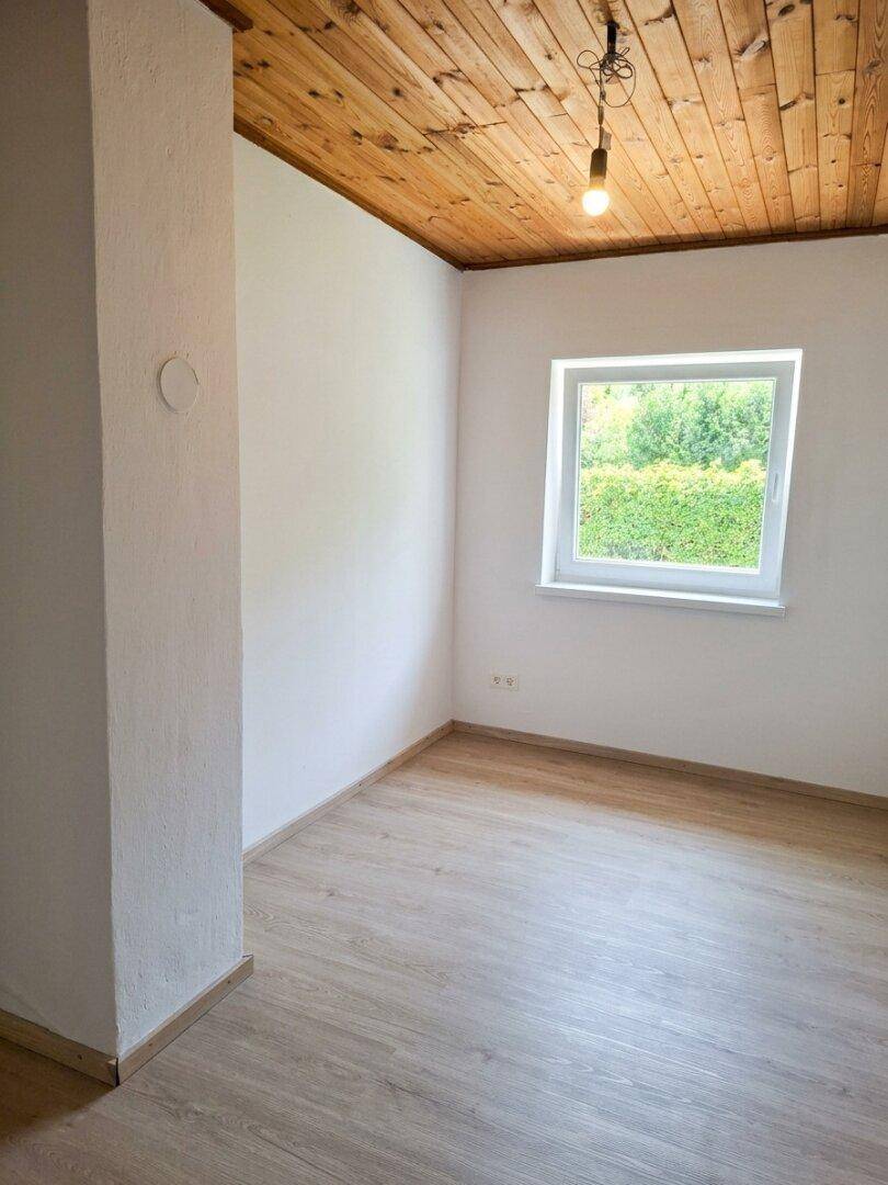 Gemütliches Schlafzimmer mit Holzbalkendecke, Laminatboden und einem Fenster mit Blick ins Grüne.