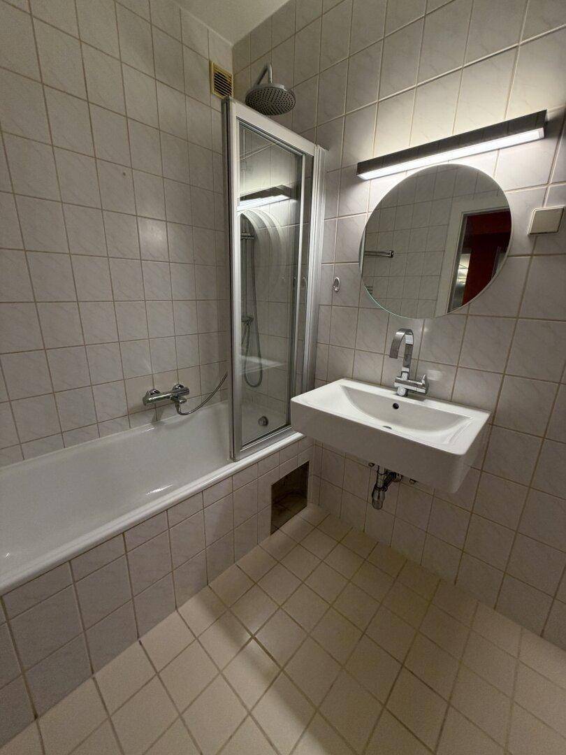 Helles Badezimmer mit weißen Fliesen, Badewanne mit Duschfunktion und modernem Waschbecken.