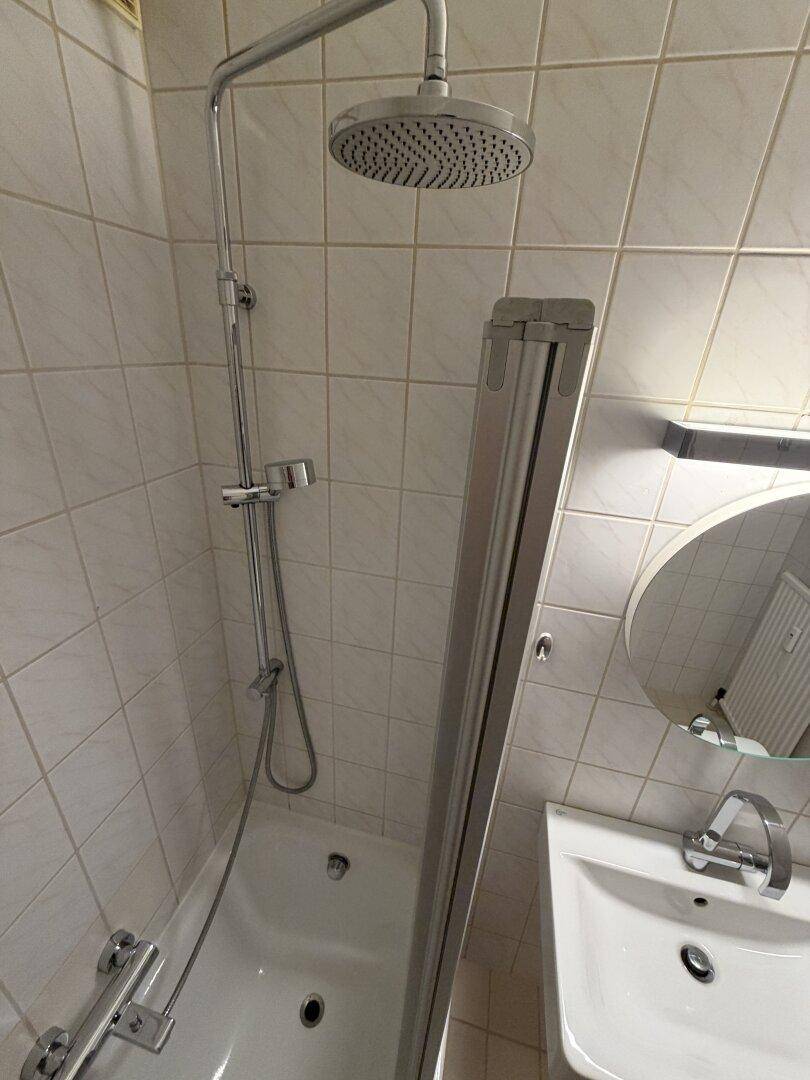 Nahaufnahme der Duscharmatur und des Duschkopfs über der Badewanne im gefliesten Badezimmer.