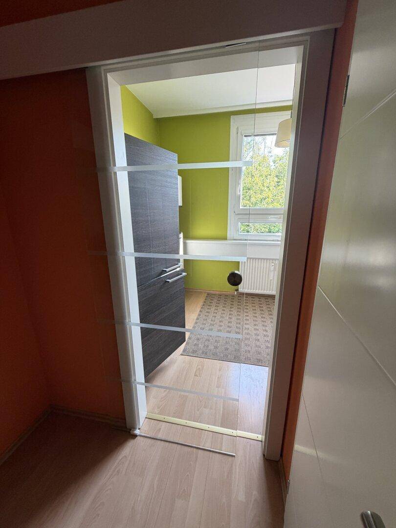 Blick durch eine moderne Tür in den angrenzenden Küchenbereich mit grünen Wänden und Fenster.