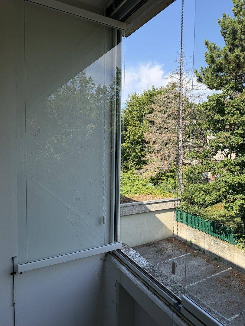 Blick von der verglasten Loggia auf die umliegenden Bäume und einen Parkplatz darunter.