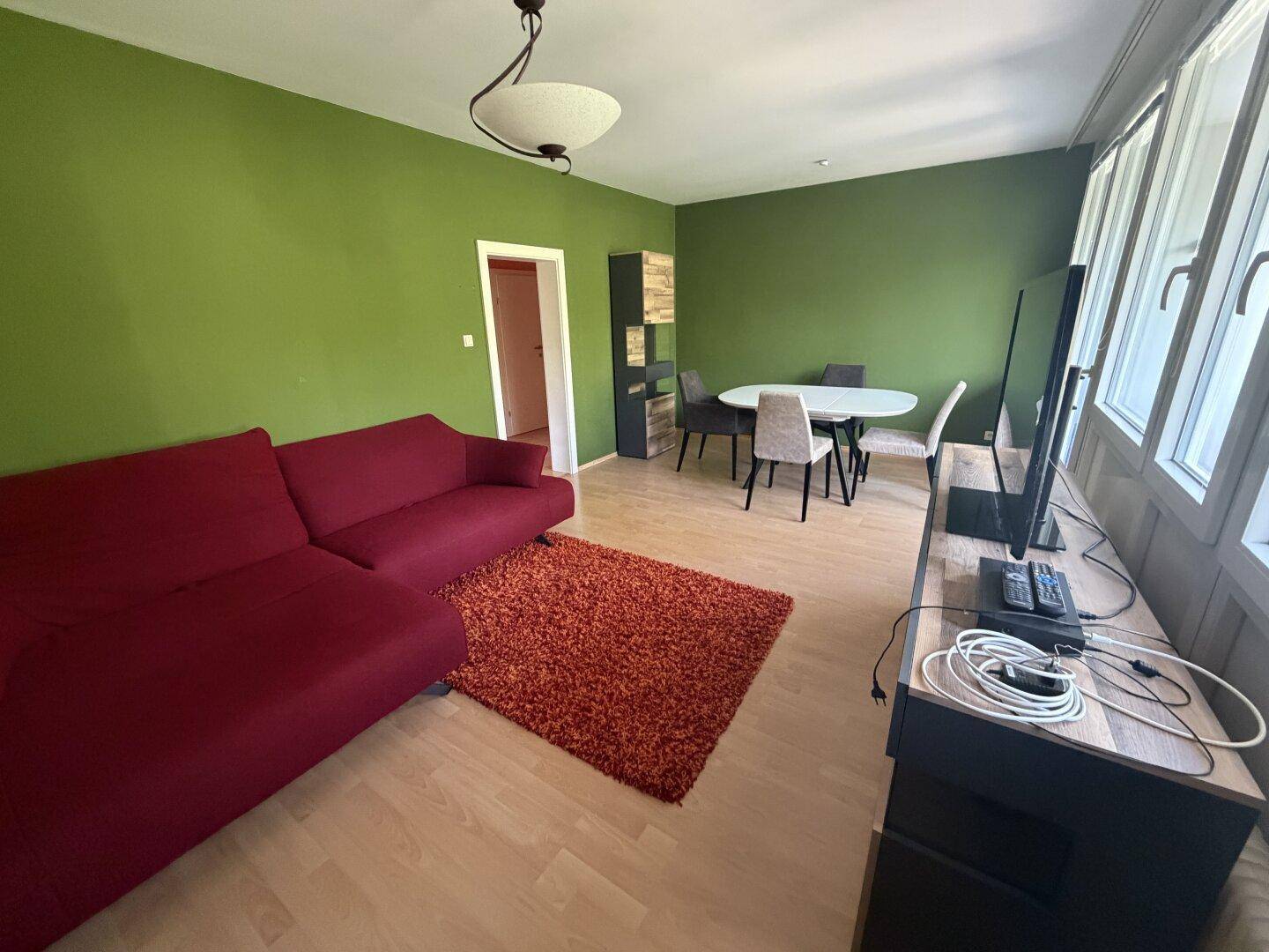 Geräumiger Wohn-Essbereich mit rotem Sofa, Esstisch und großen Fenstern, die viel Licht hereinlassen.