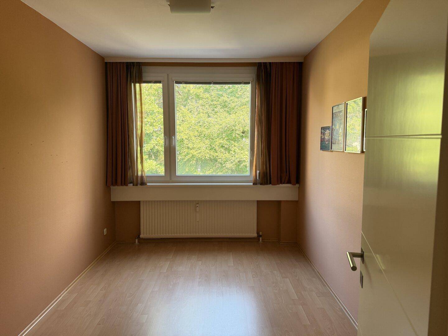Leeres Zimmer mit hellem Laminatboden, Fenster mit Vorhängen und Heizkörper, bereit zur Einrichtung.