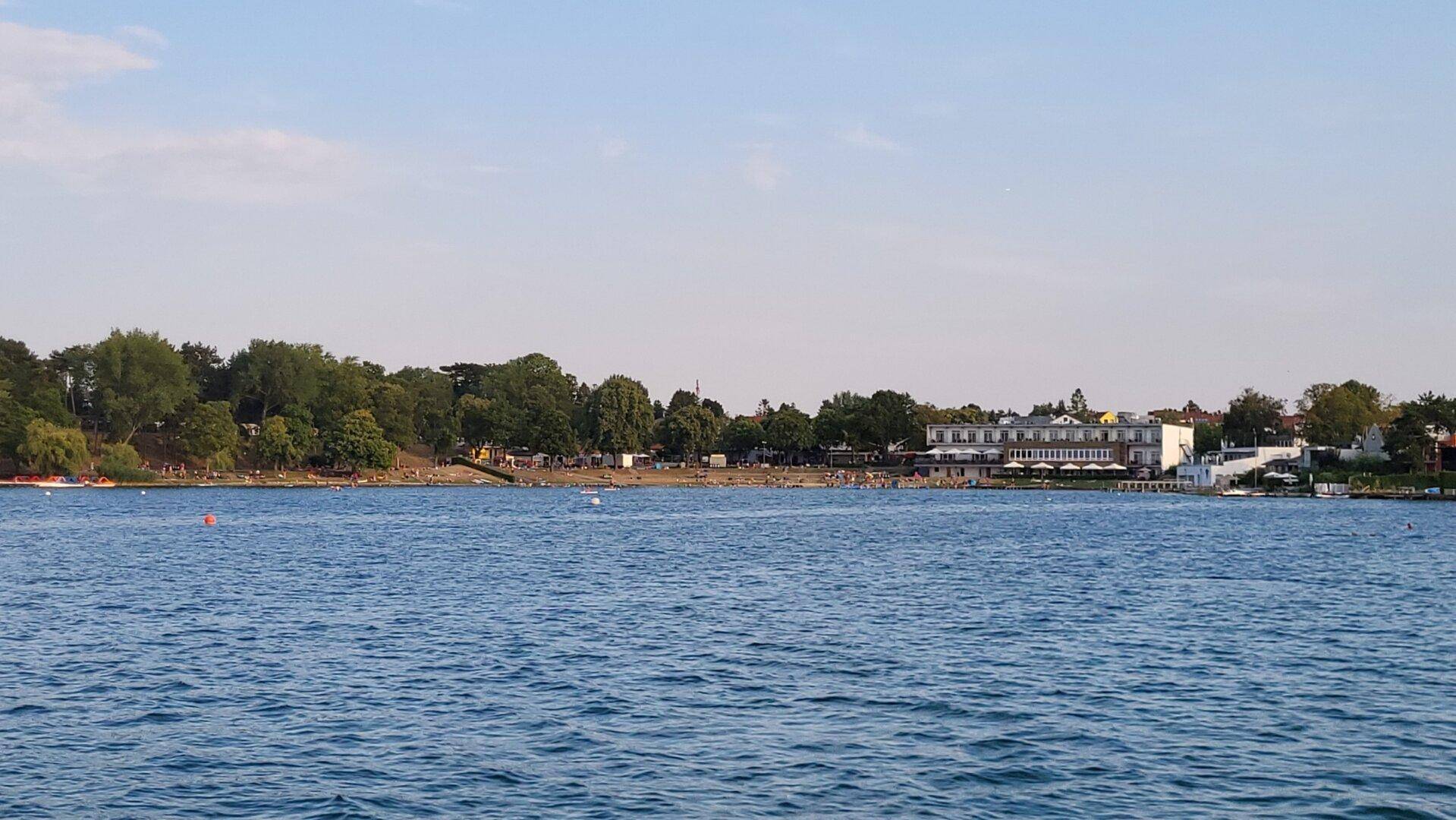Weitläufiger Blick über einen See mit bewaldetem Ufer, einem Strandbereich und einem Gebäude in der Ferne.