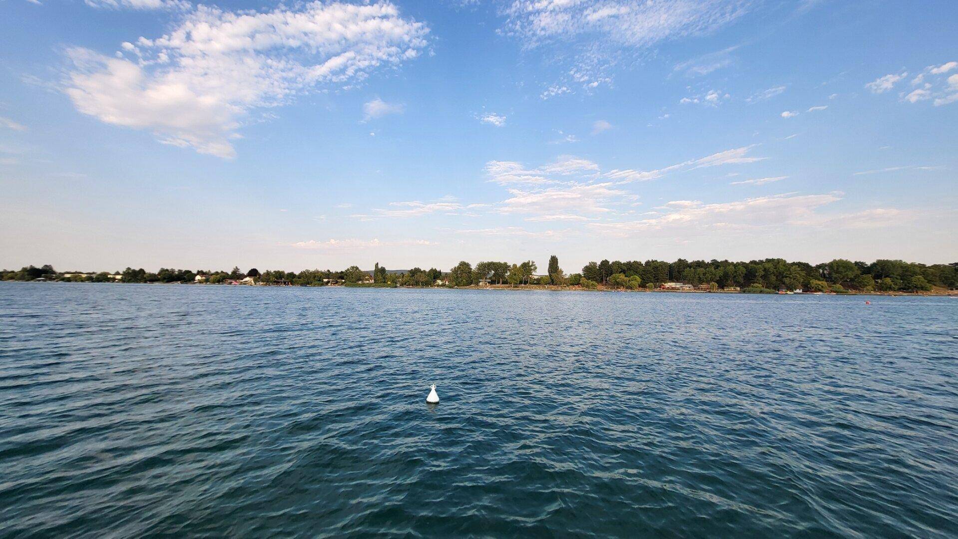 Ruhiger Ausblick auf einen See mit leicht bewölktem Himmel und einer grünen Uferlinie in der Ferne.