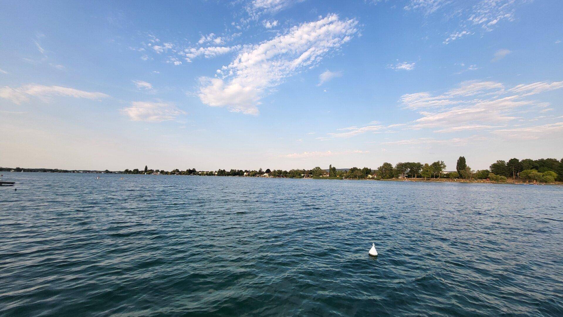 Panoramablick über einen ruhigen See unter einem weiten blauen Himmel mit vereinzelten Wolken.