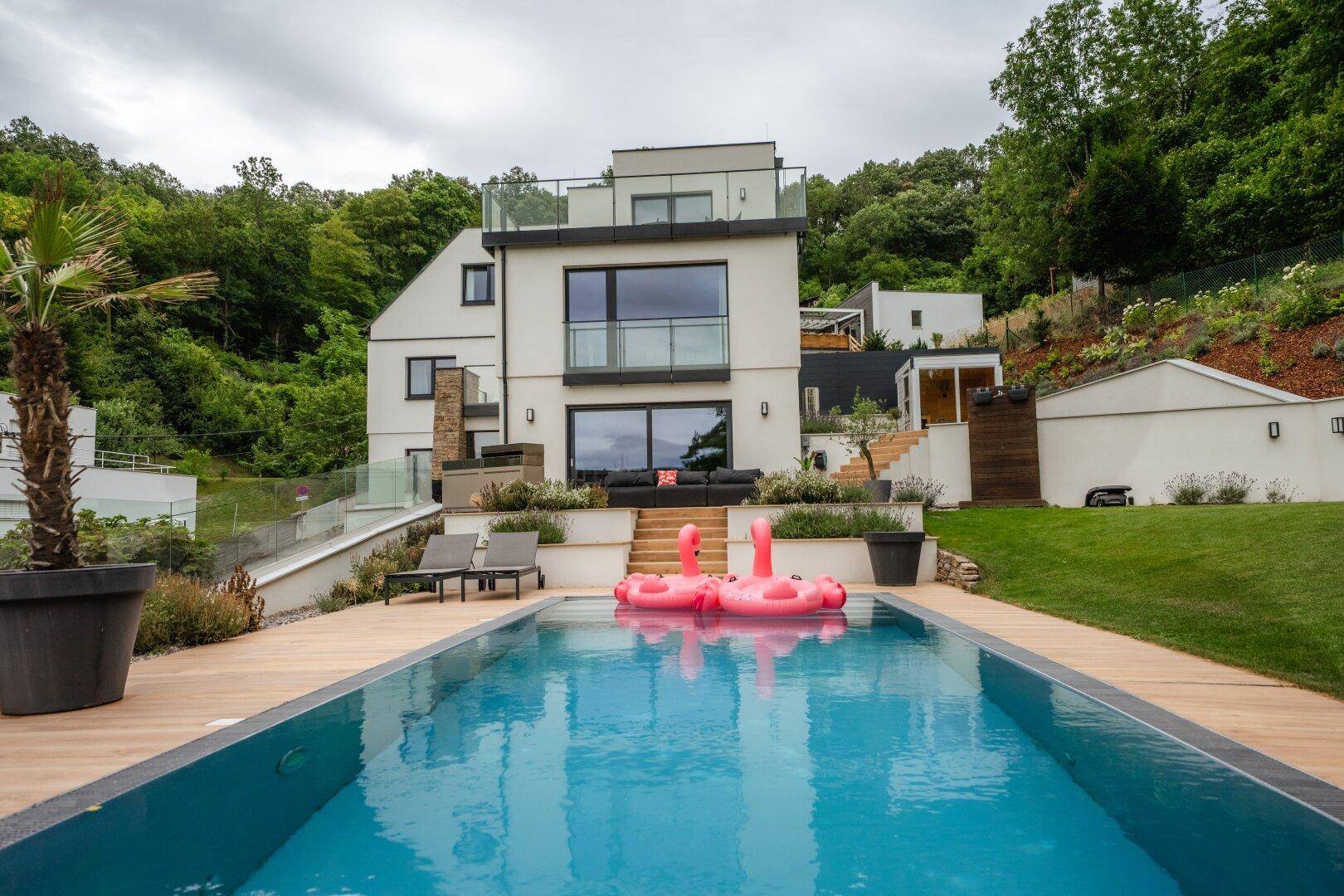 Moderne Villa mit weißer Fassade, großen Fenstern und einem einladenden Poolbereich im Vordergrund.