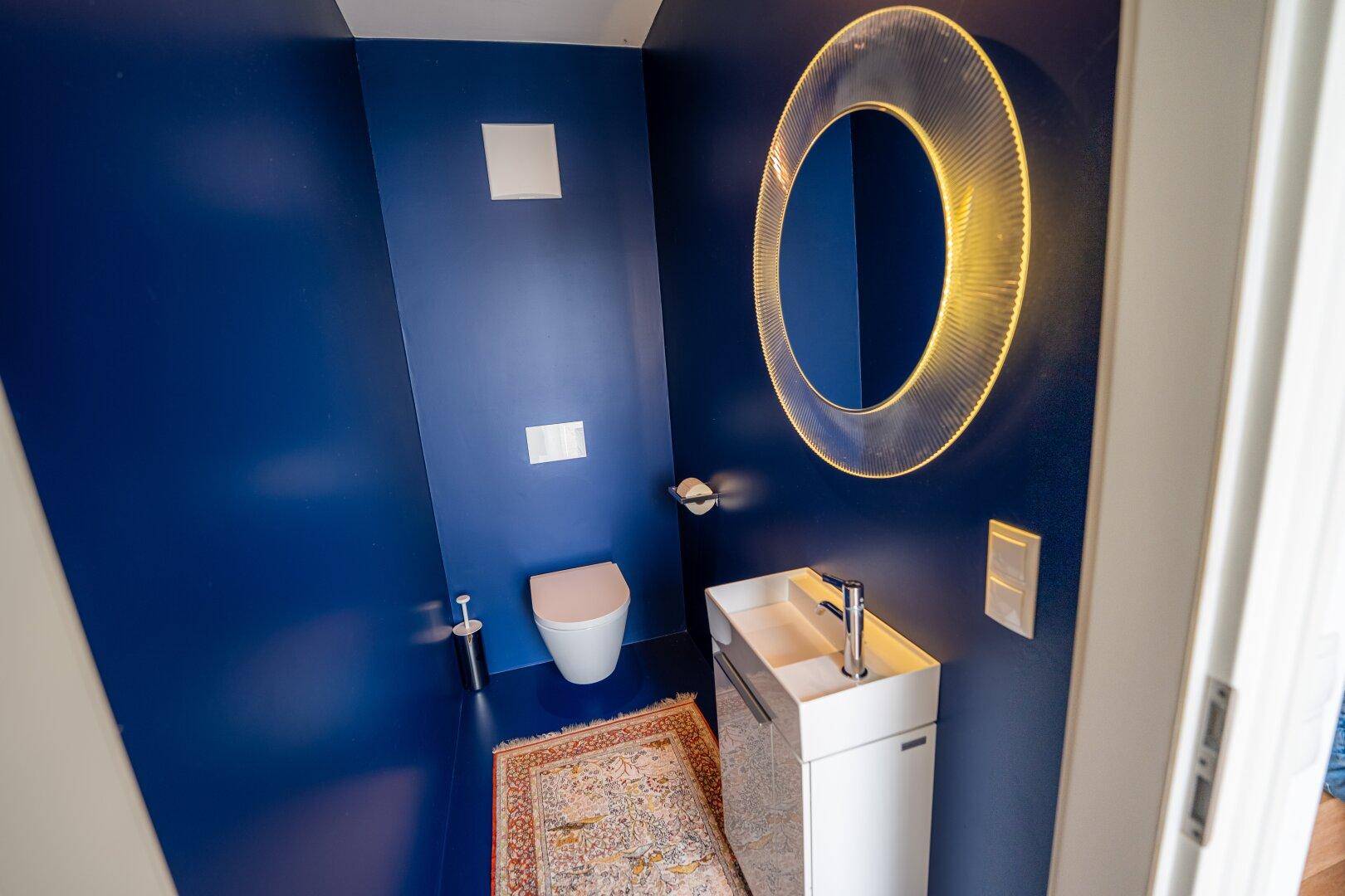 Modernes Gäste-WC mit dunkelblauen Wänden, wandhängendem WC und beleuchtetem Spiegel.
