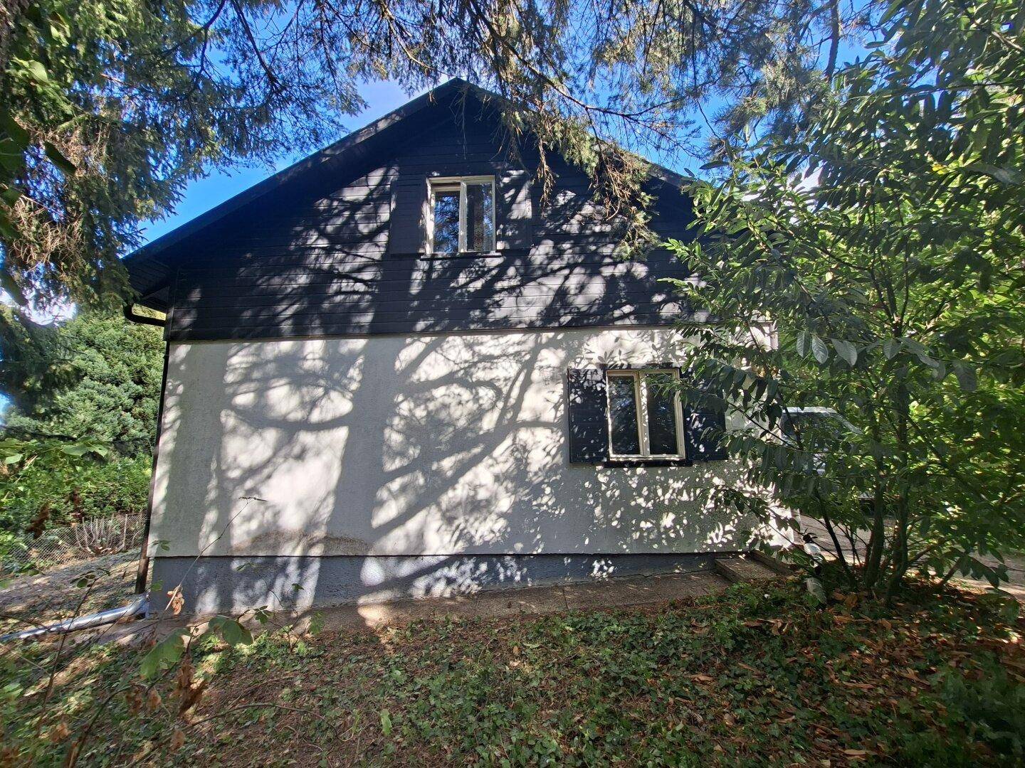 Seitenansicht des Hauses mit weißer Fassade, dunklem Dach und Schattenwurf von Bäumen.
