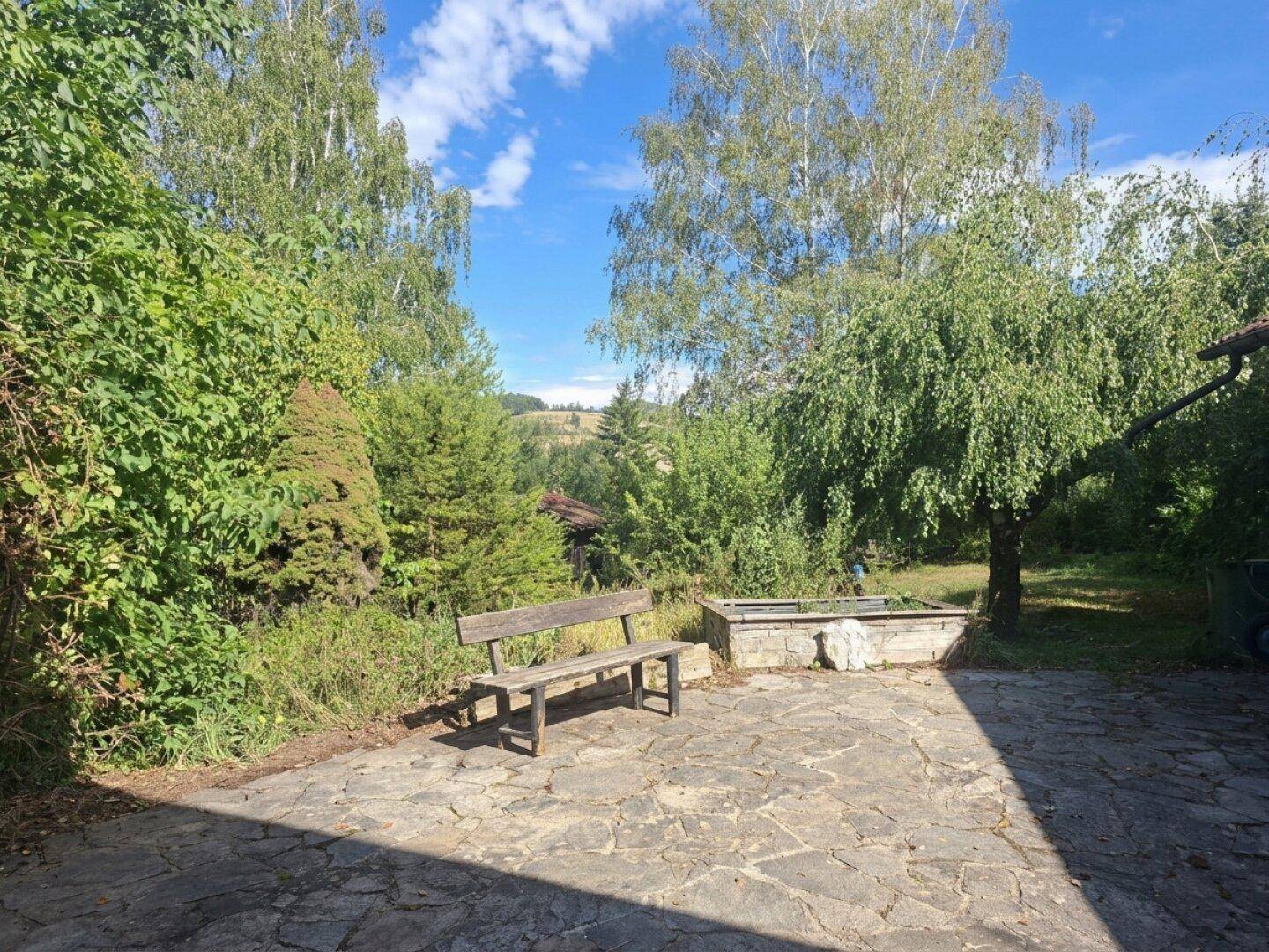 Gepflasterte Terrasse im Garten mit Holzbank und Blick auf die hügelige Landschaft.