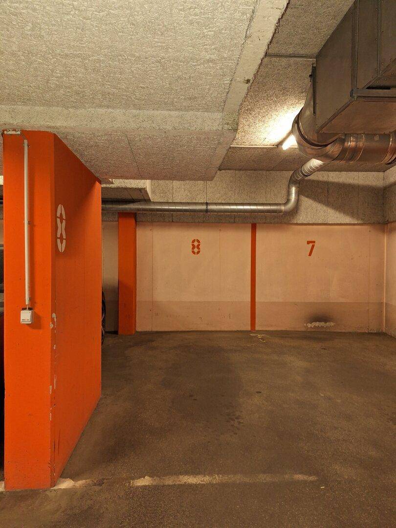 Tiefgarage mit Betonwänden, orangefarbenen Markierungen und sichtbaren Lüftungsrohren.