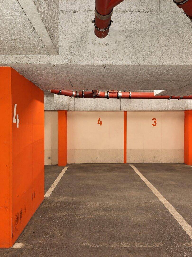 Tiefgarage mit orangefarbenen Säulen, roten Rohren und nummerierten Parkplätzen.