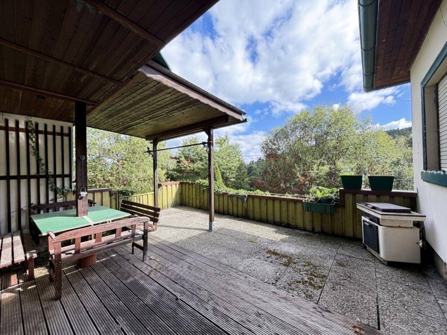 Überdachte Terrasse mit Holzdeck und Sitzgelegenheiten, umgeben von viel Grün.