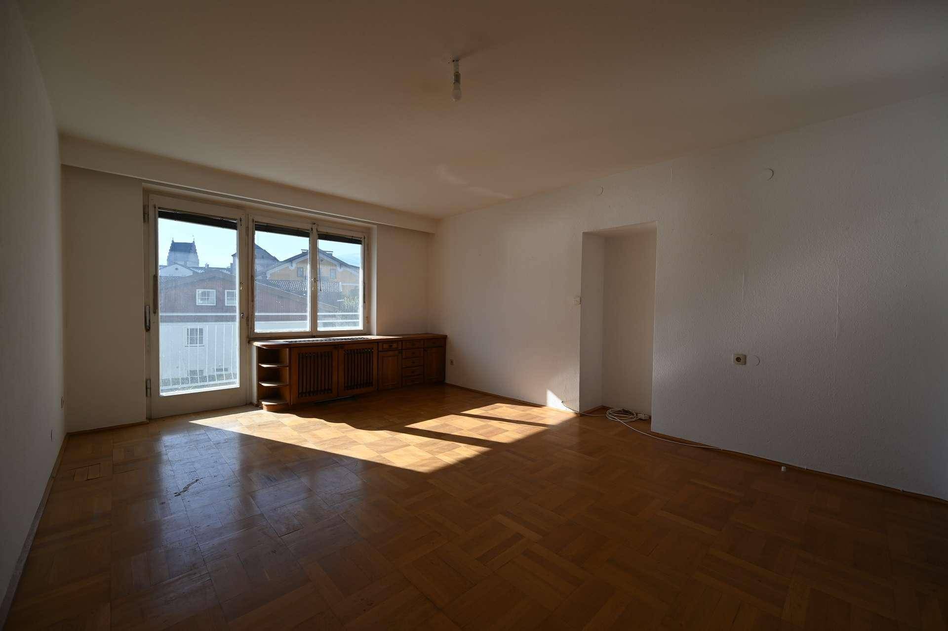 Helles Wohnzimmer mit Holzboden, großem Fenster und Zugang zu einem Balkon.