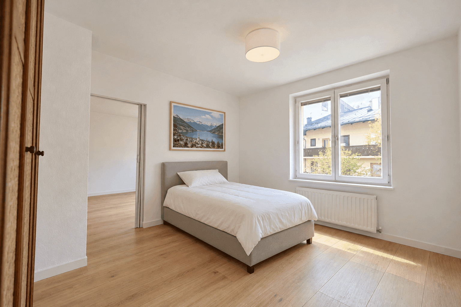 Helles Schlafzimmer mit Einzelbett, Holzboden und Blick auf die umliegenden Gebäude.