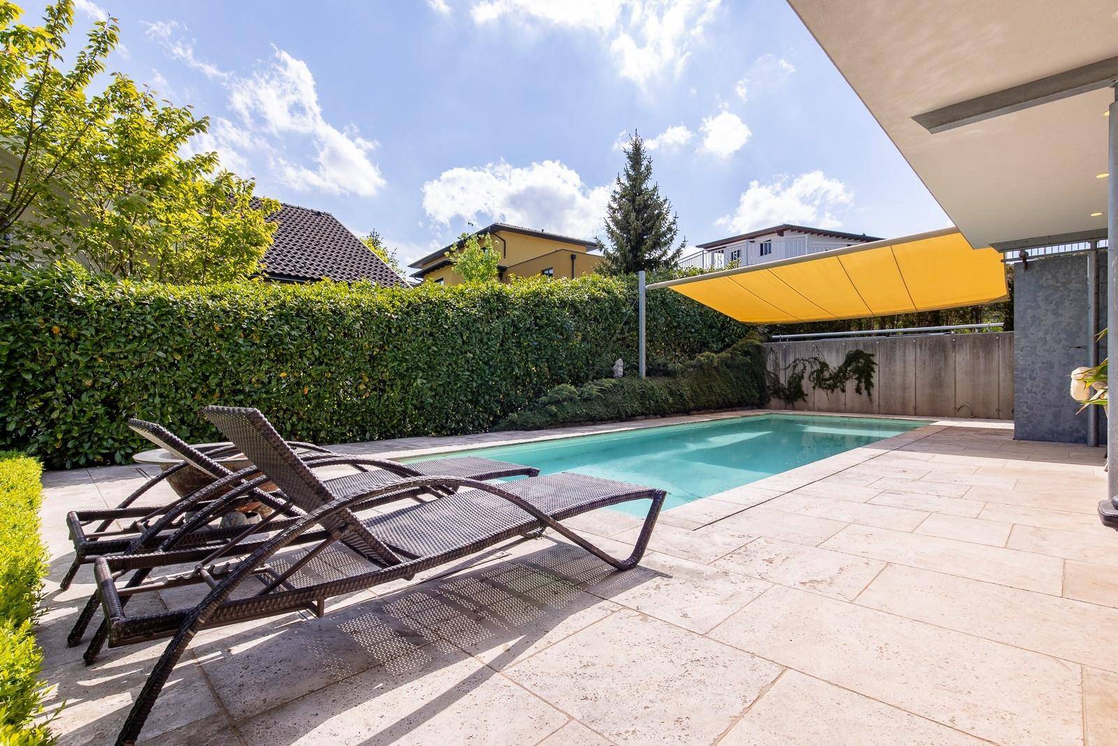 Außenpoolbereich mit Liegestühlen, Sonnensegel und gepflegter Hecke unter blauem Himmel.
