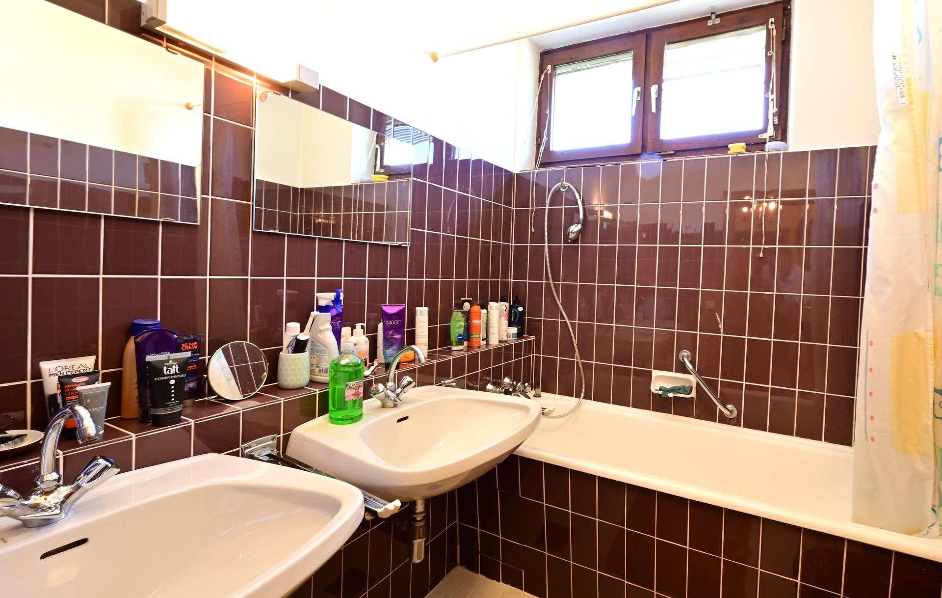Badezimmer mit braunen Fliesen, zwei Waschbecken, Badewanne mit Duschfunktion und Fenster.