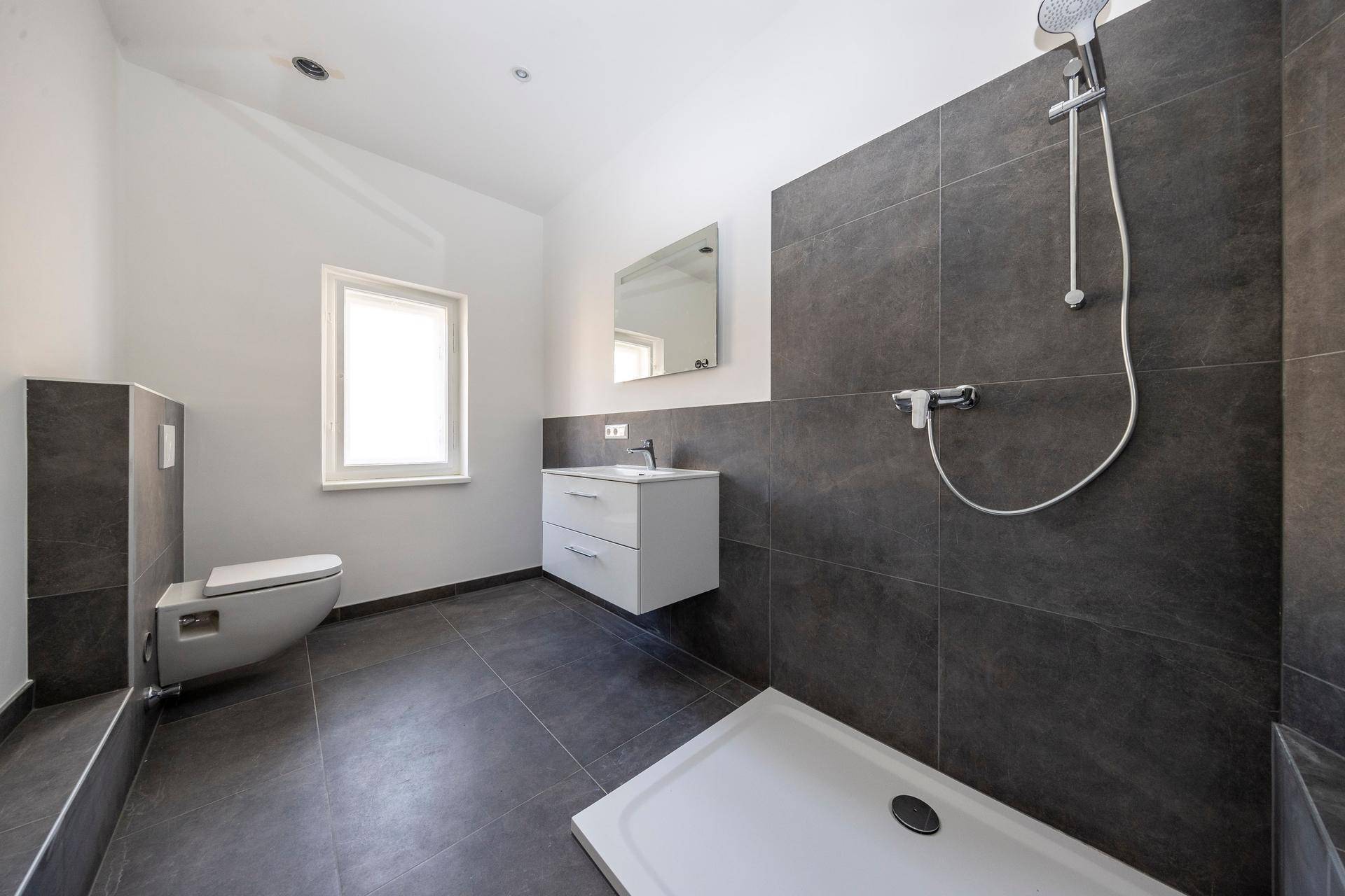 Modernes Badezimmer mit dunklen Fliesen, wandhängendem WC, Waschtisch und begehbarer Dusche.